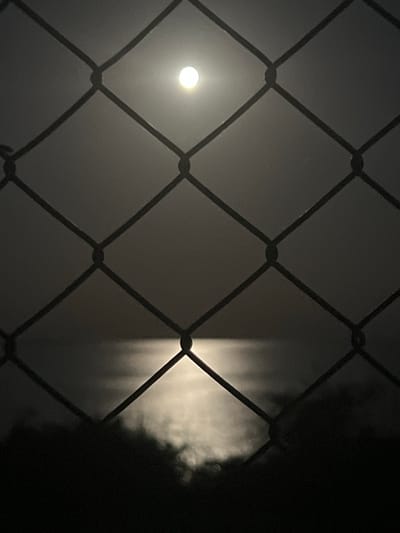 The image shows a full moon partially obscured by a chain-link fence. The moon is bright and appears to be in a hazy or foggy sky, with its reflection visible on a body of water below. The lower part of the image is dominated by dark, indistinct shapes, possibly trees or other vegetation. The overall scene is dark and atmospheric, with the fence in the foreground creating a sense of separation or observation. The location is identified as Durankulak, Bulgaria, implying it is an outdoor scene at night. There are no people or specific actions depicted.