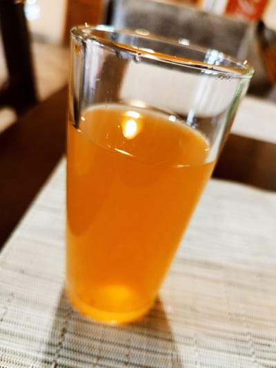A glass of orange liquid, likely juice, sits on a textured placemat. The liquid is a vibrant orange color and is catching the light, creating reflections on the surface of the glass. The glass itself is clear and appears to be made of glass, with a slightly wider opening than its base. The background is blurred and out of focus, showing hints of darker furniture and a lighter surface. The overall impression is a close-up shot of a beverage on a table.