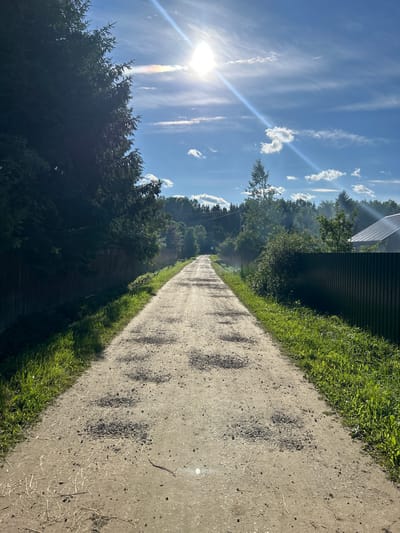 The image depicts a long, unpaved country road in the village of Слава Снт, Russia, extending into the distance under a bright, sunny sky with scattered clouds. Tall evergreen trees line the left side of the road, casting a shadow over a wooden fence. The right side of the road features a dark metal fence, overgrown with vibrant green grass. The road surface is rough and uneven, with patches of gravel and debris scattered across it. In the distance, the road appears to curve slightly, leading towards a dense forest bordering a few scattered houses. The sun is high in the sky, with its rays creating a lens flare and a faint rainbow effect in the clouds. There are no people visible in the image, and no actions are taking place. The overall scene is a serene, rural landscape bathed in the warm light of a summer day.