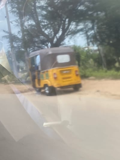 The image depicts an auto rickshaw, likely a Bajaj RE or similar model, driving on a road in Karmajiji, Nigeria. The auto rickshaw is predominantly yellow with a brown or grey roof. The background features trees and vegetation. The photo is taken through a window, possibly from inside another vehicle, as evidenced by the blurry reflection and white interior panel.