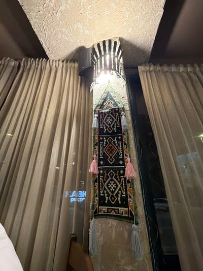 The image shows a decorative tapestry hanging from a pillar in what appears to be an indoor establishment. The tapestry is dark with intricate patterns in white, orange, and green, and features several tassels, two of which are pink and two are light gray. Above the tapestry, a light fixture with vertical slats illuminates the area. A dark gray speaker is mounted on the pillar above the light. The pillar itself is textured, resembling rough stone. To the left and right of the pillar are large, pleated curtains, light beige in color, with a subtle shimmer. Through a window or opening behind the tapestry and curtains, a reflection of blue neon lights spelling out "HABK" and "РИСТО" is visible, suggesting the presence of a sign from across the street or within the establishment. A portion of a white garment and a wooden chair are visible in the lower left and lower right of the frame, respectively. The ceiling above the pillar is also textured and light beige. The scene is indoors, with artificial lighting. The location is identified as Kumbor, Montenegro.