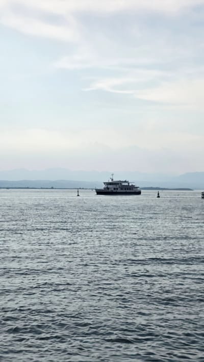 This media file depicts a ferry boat on a body of water, likely a lake or a wide river, with a mountainous landscape visible in the distance. The water is choppy, creating a textured surface with many small waves. The sky is bright and appears to be a clear day with some wispy clouds, suggesting it is daytime.

The ferry is a dark, multi-deck vessel with a relatively flat profile. It is positioned in the middle ground of the image, appearing to be in transit. Two buoys are visible in the water, one closer to the ferry and another further in the foreground, indicating a marked channel. The distant shore is characterized by a hazy, blue-toned mountain range. The overall impression is one of a serene journey across a body of water, under a bright sky, with a backdrop of natural scenery. There is no visible text in the image.