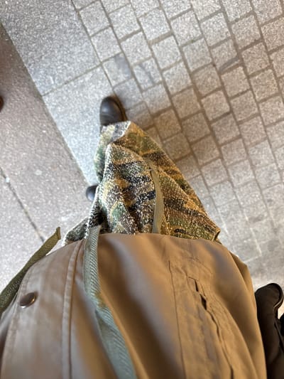 The image shows an overhead view of a person standing on a gray brick-paved street. The person is wearing a tan jacket with a visible pocket and a military-style camouflage skirt. The person is wearing black shoes. The location cannot be determined to be Palaiseau, France based on the image provided.
