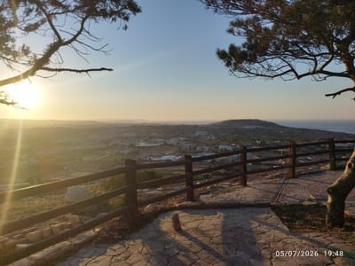 The image depicts a scenic overlook in Għargħur, Malta, at sunset. The foreground features a paved walkway with irregular stone slabs and a wooden railing bordering a sloped area. A weathered tree trunk is visible on the right side of the frame. Beyond the railing, a town stretches out towards the coast, with buildings and roads nestled amongst hills. The sun, low on the horizon and casting long shadows, illuminates the scene with a warm, golden light. The sky transitions from a pale yellow near the horizon to a clear blue overhead. The date and time stamp "05/07/2026 19:48" is visible in the bottom right corner. There are no people visible in the image.