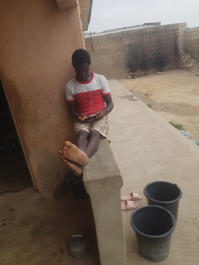 A man is reclining on a concrete structure outdoors in what appears to be Dutsinma B, Nigeria. He is wearing a red and white shirt, patterned shorts, and has earphones plugged in. He is looking at a phone he is holding. Near him are two black buckets, a pair of pink sandals, and a small metal container. A brick building is in the background, and the sky is overcast.