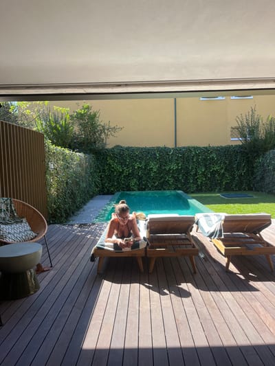 An overhead view captures an outdoor residential patio in Lordelo do Ouro e Massarelos, Portugal, featuring a swimming pool and a sunbathing area. The foreground displays dark composite wood decking. On the left, a wicker armchair with a green and white patterned cushion and blanket sits next to a cylindrical concrete side table. In the mid-ground, a female individual, wearing a dark bikini and eyeglasses with her hair in a bun, lies prone on her stomach on a wooden sun lounger, holding and looking at a smartphone. A white and dark green striped towel is draped over her lounger. Two additional empty wooden sun loungers, one with a similar striped towel, are positioned to her right. Beyond the loungers, a rectangular swimming pool with turquoise water occupies the center-rear. The pool is bordered by green, dense ivy-covered walls or hedges. Behind the vegetation, a tall, plain yellow building wall is visible, featuring two small rectangular windows on its upper right section. Sunlight illuminates the pool and sun loungers, casting shadows on the decking. A vertical slatted privacy screen runs along the far left edge of the patio.