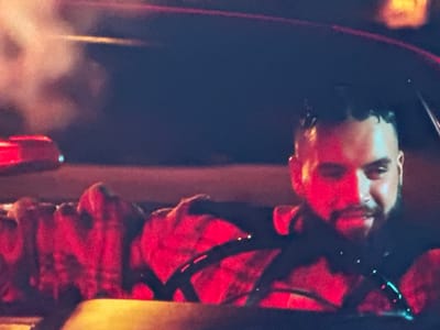 A man with a beard and short, spiky hair is in the driver's seat of a car. He is looking towards the camera with a slight smile. The interior of the car is lit by red and yellow lights. The man is wearing a dark shirt. Smoke or fog is visible in the background. The image appears to be taken at night, likely in an urban or suburban environment. The location is identified as Ravno pole, Bulgaria.