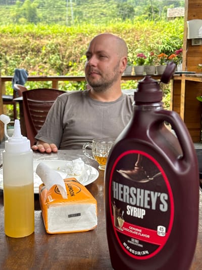 The image captures a casual outdoor or semi-outdoor scene, likely in Du Già Commune, Vietnam, featuring a man and several items on a wooden table. A bald man with a beard, wearing a grey t-shirt, is positioned at the table, looking down with a focused expression. His right hand is visible on the table.

The most prominent object is a large, brown plastic bottle of Hershey's Syrup, with its cap open, placed on the table. Its label clearly displays "HERSHEY'S SYRUP," "GENUINE CHOCOLATE FLAVOR," "FAT FREE," and "NET WT 48 OZ (3 LB) 1.36 KG," along with a "45" calorie indicator. Also on the table are a clear glass mug containing an amber liquid, a white plate, and an orange and white tissue box labeled "LAMOON" and "300系列," from which a white tissue has been pulled. A white squeeze bottle filled with a yellow liquid is also present.

The background is dominated by lush, green vegetation, indicative of a natural, perhaps mountainous or rural environment. Parts of a rustic wooden structure are visible, suggesting an open-air dining area. The bright lighting suggests it is daytime. The man appears to be engaged in a meal or snack, with various condiments and drinks at hand in this relaxed setting.