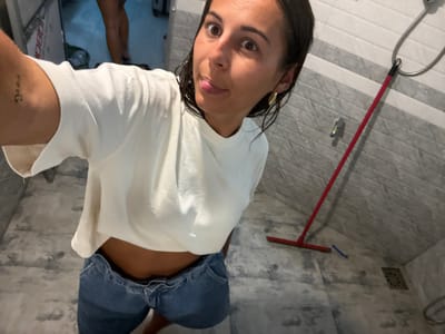 The image depicts an indoor scene in a bathroom or shower area, located within the city of Nilwella, Sri Lanka.
A female subject, appearing to be an adult, is positioned in the foreground, taking a selfie. Her dark hair is wet. She is wearing a white, short-sleeved crop top and blue denim shorts that are partially lowered, exposing her midriff. A small, cursive tattoo is visible on her upper left arm. She wears a gold-colored hoop earring in her right ear. Her facial expression shows wide eyes and a slightly extended tongue.
In the blurred background, the lower legs and feet of a second individual are partially visible.
The environment features tiled walls with alternating light and dark grey horizontal stripes. The floor is light grey with a square drain grate visible in the lower right. A shower head with a flexible hose and a silver mixer tap are mounted on the wall. A red-handled floor squeegee leans against the right wall, and a small blue object rests on the floor nearby.