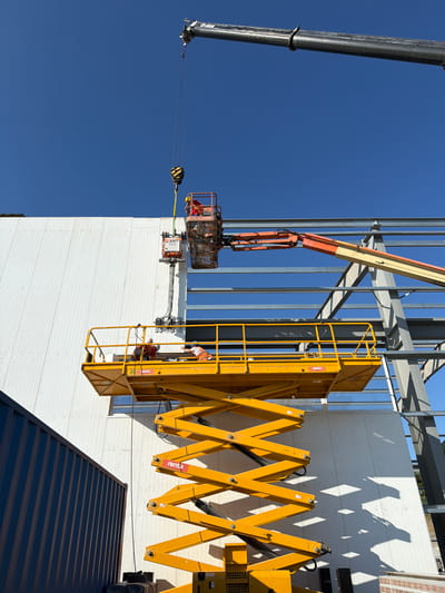 The image shows a construction site in Stróża, Poland, under a clear blue sky. A large industrial building with white siding is under construction. A dark blue shipping container is partially visible on the left.
Multiple pieces of construction equipment are in use. At the top, a crane with a hook is suspended in the air, likely in the process of lifting an object. Below the crane, a man in an orange safety vest is working in a red elevated platform, which is attached to an articulated boom lift. This boom lift, painted orange and yellow, extends towards the building.
Further down, a large yellow scissor lift is positioned in front of the building. Two construction workers are on its elevated platform. One worker, wearing an orange safety vest, is seated. The other worker, in a blue shirt and orange safety vest, is standing and appears to be interacting with the seated worker. The scissor lift has "rent" labels visible on its structure.
The building's metal frame is exposed in several areas, indicating ongoing structural work. Sunlight casts distinct shadows on the white siding of the building. The scene depicts an active construction project with workers utilizing heavy machinery to advance the building process.