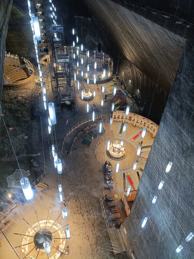 This is an overhead, high-angle shot of an underground amusement area within a salt mine. The cavernous space is illuminated by numerous cylindrical pendant lights, some of which appear to emit a soft blue glow, while others are a warmer white. The rough, textured walls of the salt mine are visible on the right and left sides of the frame, with striations and mineral deposits evident.
The scene is populated by a variety of attractions and activities. In the center-left, there is a large amphitheater-like seating area carved into the mine. Further into the mine, a complex metal structure, possibly an elevator or a ride, is visible, along with more seating areas and hanging lights.
Scattered throughout the open floor of the mine are several recreational areas. There's a mini-golf course with brightly colored holes and pathways. A carousel is situated in the middle ground, surrounded by people. A playground with slides and climbing structures is also visible in the upper portion of the frame. Several groups of people are seen walking, sitting at tables, and engaging in the activities.
The ground of the mine appears to be covered in a sandy or gravel-like material, with wooden pathways and barriers marking out different zones. The overall impression is one of a vibrant and unique entertainment venue carved into the natural formations of a salt mine. The time of day is indeterminate, but the artificial lighting suggests it is either evening or the mine is illuminated for continuous operation. No specific weather conditions can be inferred from the indoor setting. The location is identified as Turda, Romania. There is some faint, unreadable text on a banner on the metal structure in the upper left portion of the image.