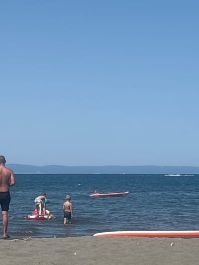 The image depicts a bright, sunny day at a beach, with clear blue skies and calm, dark blue water. In the foreground, a person's legs are visible, standing on a sandy beach. The middle ground shows several individuals in the water. Two young boys are standing near each other in the water. One boy is on a red inflatable raft, with another child lying on the raft as well. Another young boy stands to the right of them, also in the water. Further out in the water, a red inflatable raft is floating, with a person on it. In the background, a low, hazy mountain range is visible in the distance, bordering the water. The overall scene suggests a leisurely day at the beach.