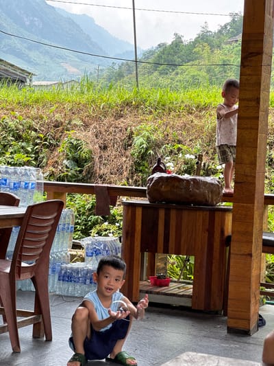 This outdoor image, taken in Du Già Commune, Vietnam, depicts two young boys on a semi-covered patio or balcony overlooking a lush, mountainous landscape. The sky above the mountains appears overcast or hazy, suggesting a cloudy day.

In the foreground, a young boy with dark hair is crouched on a grey concrete floor, looking directly at the camera with a slight smile. He wears a light blue t-shirt and dark blue shorts, with dark green open-toed sandals on his feet. His hands are raised as if playing with small, glistening droplets, possibly bubbles or water. To his left, behind him, are stacks of clear plastic bottled water. A brown plastic chair is partially visible in the bottom right corner.

Further back and to the left, another young boy stands, peering over a wooden beam of the structure. He wears a light-colored sleeveless top and patterned shorts, with bare feet visible. He appears to be looking out towards the scenery. On his shorts, fragmented text "OID ME" is faintly visible.

The setting features robust wooden beams and railings, suggesting a rustic yet sturdy construction. On a wooden surface between the two boys, there's a large, dark, irregularly shaped object resembling a stone or carved wood. Below the wooden structure, a red plastic basket and a pair of dark blue flip-flops are visible. Beyond the patio, abundant green grass and foliage cover a sloping hillside, leading to layers of misty, verdant mountains in the distance. A dark power line stretches across the middle ground of the scenic backdrop, with small cultivated patches visible on some hillsides. The overall scene conveys a sense of casual play and observation in a tranquil, natural environment.