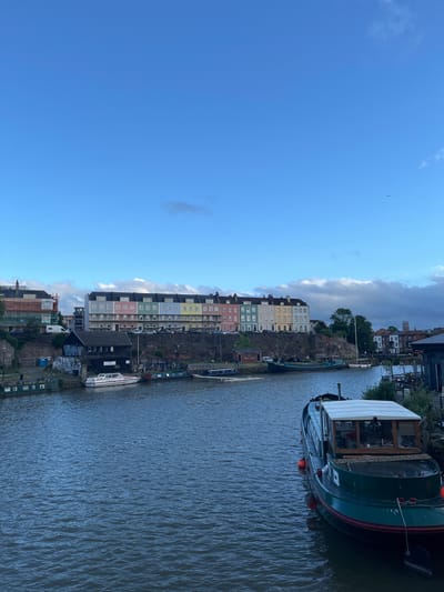 The image depicts a vibrant waterside urban scene, likely a river or canal, under a bright blue sky with scattered white and grey clouds. The main subjects are a long row of colorful, multi-story buildings lining the left bank of the waterway, and several boats docked on the water.
In the foreground and mid-ground on the right, a prominent dark green canal boat with a white-canopied cabin is moored, featuring red buoys along its side. Other smaller boats are visible further down the waterway. The water itself shows ripples and reflections, suggesting a gentle flow.
Along the left bank, a distinctive architectural feature is a series of pastel-colored buildings (including shades of pink, yellow, light blue, and mint green) built into or against a rocky embankment, which also features some greenery and trees. Further down the bank (towards the left), additional buildings, some appearing older or industrial, and some construction scaffolding are visible.
The scene conveys a peaceful daytime atmosphere. The time of day appears to be during good daylight hours, indicated by the clear sky and the way light illuminates the buildings. The weather is fair with partial cloud cover. A small bird can be seen soaring high in the blue sky on the upper left side of the frame. No discernible human figures or explicit activities other than the stationary boats are visible. Given the context, this scene is characteristic of areas like Bristol's Harbourside. There is no clearly legible text visible on any of the structures or boats.