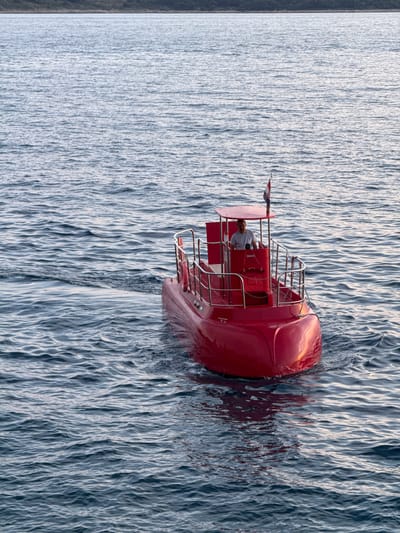 The image shows a bright red, small passenger submarine, or semi-submarine, with a clear canopy, navigating through calm, blue-grey water. A man, wearing a light-colored t-shirt, is visible at the helm of the submarine, steering the vessel. A flag, appearing to be the flag of Croatia, is planted on the rear of the submarine's roof. The submarine is creating a wake as it moves. In the background, a dark, low-lying coastline with vegetation is visible on the horizon. The lighting suggests it is either early morning or late afternoon, with a soft, warm light reflecting on the water and the submarine. This scene is characteristic of the waters surrounding Primošten, Croatia, a coastal town known for its clear waters and tourist activities.