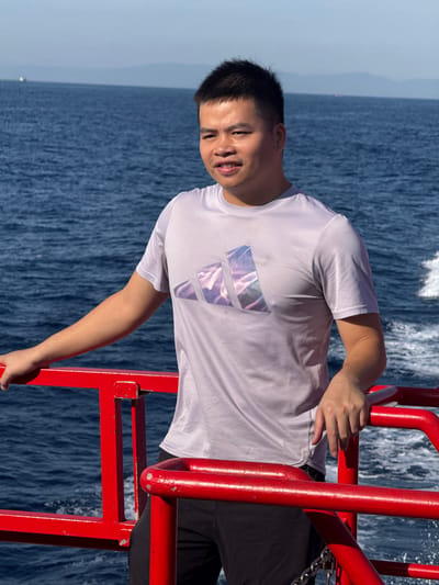 A man stands on the deck of a boat, leaning on a red railing. He is wearing a light purple t-shirt with a colorful Adidas logo and black shorts. The boat is on the water, with the sea stretching out behind him. The sky is clear and blue. The man is looking slightly to his right, with a neutral expression. The water is calm, with some ripples and a wake trailing behind the boat. In the distance, a hazy shoreline can be seen. There are no clear indications that this image was taken in Dong Son, Vietnam; the environment depicted is consistent with many coastal areas.
