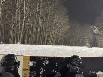 A snowboarder's perspective at night on a snowy slope surrounded by bare trees. The snowboard, with bindings secured, fills the foreground. Snow covers the ground, leading up to a line of dense trees with their branches bare against the dark sky. Ski lift support structures are present on the side of the hill.