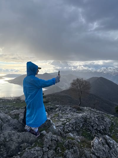 A sole individual stands on a rocky, uneven mountain ridge at an elevated position in Bogdašići, Montenegro, overlooking a vast panoramic landscape. The person, viewed from a slightly elevated rear-left angle, wears a bright blue hooded poncho, a yellow baseball cap, tan pants, white socks, and purple hiking shoes. A black backpack is partially visible behind them. They are actively holding a black smartphone vertically with both hands, extended forward, seemingly capturing the expansive view. To the left, a large, calm body of water, resembling a bay or fjord, reflects bright sunlight breaking through an overcast sky. Coastal settlements are visible along its shores. The midground and background consist of multiple layers of dark, rugged mountains. A solitary, bare deciduous tree with a contorted trunk stands on a ridge to the right of the individual. Distant mountain peaks in the far background are snow-capped under a heavy, dark grey cloud cover. The foreground is composed of large, grey rocks with sparse green vegetation and white lichen patches.