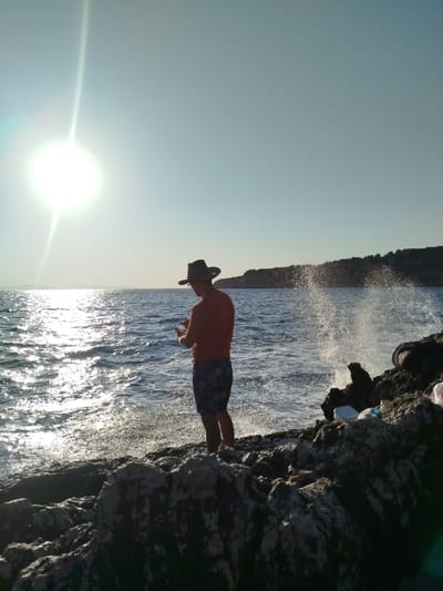 A person stands on rocky terrain by the sea in Karaburun, Turkey, facing away from the camera and towards the water. The person is wearing a hat, an orange t-shirt, and patterned shorts. They are holding a mobile device in their hands and appear to be looking at it. The sun is bright and low in the sky, casting a strong glare on the water. Waves are crashing against the rocks, creating a spray. The background shows a coastline with trees.