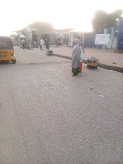 This image captures a street scene in Dakata, Nigeria, characterized by daily activity under bright, hazy sunlight, suggesting either early morning or late afternoon.
The foreground is dominated by a gray asphalt road. Two concrete-filled tire barriers, painted with yellow and black stripes, are positioned on the right side of the road, acting as dividers or bollards. A similar barrier is visible further back on the left.
In the midground, several people are present. Two women stand on the right, with their backs to the camera; one wears a light-colored head covering and garment over a green patterned skirt, and the other has a light green top with orange trousers. Further back, a small gathering of people congregates near a row of shops. One individual in a light green shirt is seen walking on the left side of the street. A yellow motorized tricycle (auto-rickshaw) is parked on the far left.
The background shows a commercial area on the left, with various shops or market stalls and more people moving about. On the right, a blue gate leads to a building, likely a public or community facility, where a large white sign is mounted. Clearly visible text on this sign reads "DANGE LOCAL GOVERNMENT COUNCIL," indicating a local administrative presence. Other text on the sign is illegible due to distance and resolution, but pertains to official information. Faintly visible text on a shop building in the left background appears to include "CONTAINER." The scene conveys a typical day in a bustling local street environment.