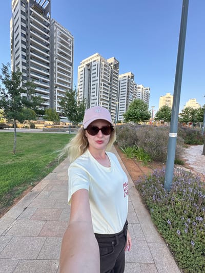 A woman is taking a selfie in an urban park setting. She is wearing a light yellow t-shirt with red text, a pink baseball cap, and sunglasses. In the background, there are modern high-rise apartment buildings and a clear blue sky. The foreground shows a paved walkway next to a grassy area with some flowering bushes and trees. The sun appears to be low in the sky, casting long shadows, suggesting it is either early morning or late afternoon. The overall mood is casual and relaxed. The red text on the woman's shirt is visible and appears to read "May NEED..." followed by an illegible word.