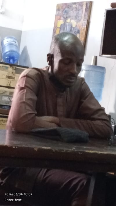 An indoor scene in the city of Dakata, Nigeria, depicts an adult male seated at a dark, worn wooden table. The man has a shaved head and wears a brown, long-sleeved, collared tunic and dark trousers. His hands are clasped on the table, and his gaze is directed downwards. A dark fabric object rests on the table in front of him. The background features light-colored walls. To the left, an inverted clear blue plastic water dispenser bottle sits atop a multi-drawer storage unit. In the center-right, a rectangular framed artwork with stylized yellow and blue figures hangs on the wall. A dark flat-screen monitor or television is partially visible to the right of the artwork, with another partially visible clear blue plastic water dispenser bottle behind it. A digital timestamp "2026/03/04 10:07" is visible in the bottom-left corner of the image.