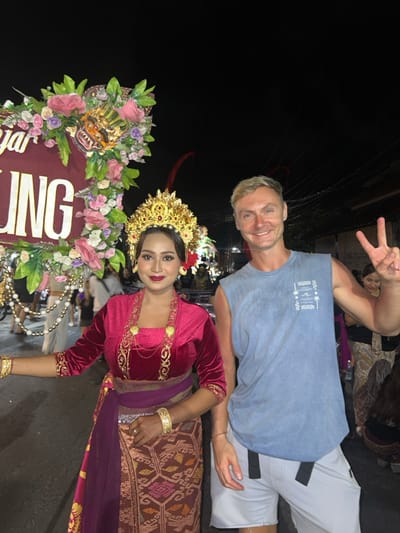 This is a nighttime photograph taken outdoors in Munggu, Indonesia, depicting two individuals posing in the foreground of what appears to be a street festival or parade.
On the left, a woman of Indonesian ethnicity smiles, looking directly at the camera. She wears traditional Balinese attire, including an elaborate golden crown, a long-sleeved deep red velvet top, a multi-layered golden necklace with round pendants, matching golden bracelets on both wrists, and a patterned woven sarong in brown, orange, and purple hues secured with a purple sash. Her left arm is slightly extended to her side.
On the right, a Caucasian man with light-colored hair smiles, also looking directly at the camera. He wears a light blue sleeveless t-shirt with a white graphic design on the left chest and light grey shorts with black strap accents. His right hand is raised, forming a "peace" sign.
Between and behind the two individuals, partially visible on the left, is a large, ornate burgundy structure, possibly part of a float or banner. It features white lettering, with "NG" clearly visible. This structure is heavily decorated with a vibrant arrangement of pink, purple, and green artificial flowers, and centrally features a golden, stylized mask or head resembling a mythical creature, with prominent fangs and red accents. A string of white spherical beads hangs from the decoration.
The background is dark, indicating night, with artificial lighting illuminating the scene. Other blurry figures are visible in the mid-ground, some appearing to wear traditional garments, suggesting a crowd gathered for an event. The ground surface appears to be paved.
