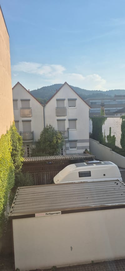 The image captures a view of residential buildings in Baden-Baden, Germany, under a clear blue sky with scattered clouds. In the foreground, a white vehicle with solar panels on its roof is parked, likely a camper or a similar recreational vehicle. This vehicle is situated on top of a corrugated metal roof structure, possibly a garage or shed. To the left of the vehicle, a building with light-colored walls and red-tiled roofs is visible. This building features multiple windows, some of which are covered by roller shutters, and small balconies. Lush green ivy climbs the side of this building. Behind these houses, a larger, modern building with horizontal window bands and a darker facade can be seen. Further in the background, a tree-covered hill dominates the landscape, topped by a distant tower. The scene is static and appears to be taken from an elevated perspective, possibly a window.