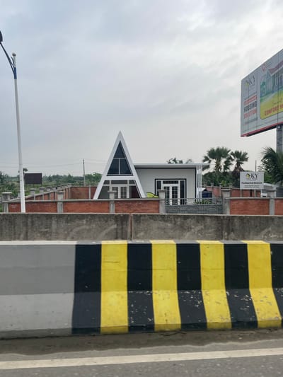 The image depicts a building under construction or recently completed in Purbachal, Bangladesh. A sign indicates it's the "Purbachal Branch" of "KY TWO TONE," a company offering structured housing solutions. The architecture includes a distinctive A-frame component and a white, rectangular section. Adjacent to the building are brick-lined structures, potentially individual housing units or part of a larger development. In the background is greenery. The image also shows a black and yellow striped barrier. A billboard advertises the same company and their housing solutions.