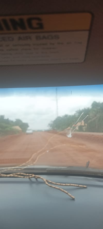 The image shows a view from inside a vehicle, looking out through the windshield. The windshield is partially obscured by splatter. The road ahead appears to be unpaved. Green vegetation flanks the road. A warning label is visible at the top of the image. A small, forked branch rests on the dashboard. The location is Oto, Nigeria.