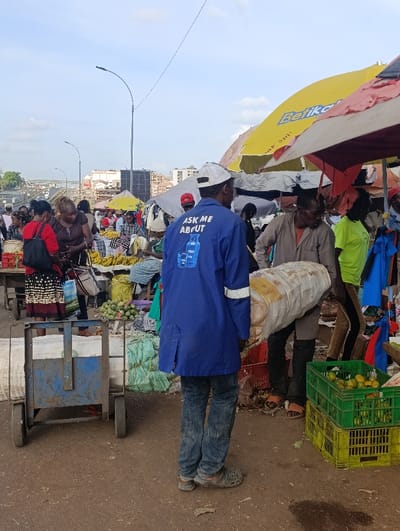 This image depicts an active outdoor market situated in Kiuu ward, Kenya, under a partially cloudy sky. The ground surface is primarily unpaved earth.
In the immediate foreground, a male subject with his back to the viewer stands slightly right of center. He wears a blue long-sleeved work shirt with the phrase "ASK ME ABOUT" above a depiction of a gas cylinder, followed by "GAS" printed vertically below it. He wears blue jeans, grey Crocs, and a white baseball cap. To his left, a blue, four-wheeled handcart is partially visible.
To the right of the central subject, another male individual, wearing a brown shirt and dark pants, is bent forward, carrying a large, white, cylindrical sack over his right shoulder. He wears orange sandals. Behind him, a third individual in a bright green shirt is partially visible. Adjacent to these individuals, stacked green and yellow plastic crates hold numerous oranges or similar round fruits.
Throughout the midground, multiple market stalls are visible, characterized by large, colorful umbrellas providing shade. A prominent yellow umbrella displays the word "Betika." Other umbrellas are red and white. Under these umbrellas, various goods are displayed, including bundles of green produce, clothing, and bananas arranged on tables. Several individuals, primarily women, are engaged in market activities; one woman in a red shirt and a black-and-white patterned skirt carries a black shoulder bag near a produce stall displaying bananas.
In the background, beyond the immediate market area, multi-story urban buildings are visible, along with a multi-lane highway or bridge structure with vehicles. Streetlights extend upwards against the light blue and white sky. Approximately 15-20 people are discernible within the frame, participating in the market environment.