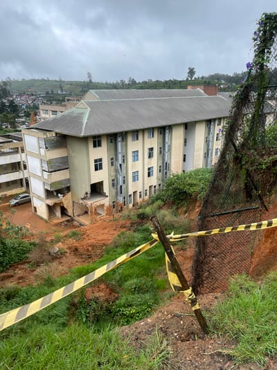 The image depicts a building in Nuwara Eliya, Sri Lanka, that has sustained significant damage, likely due to a landslide. The building's facade shows collapsed sections, exposing the interior structure. A silver car is parked in front of the building. Yellow and black caution tape is visible near the edge of the damaged area, suggesting a hazardous zone. The surrounding environment includes foliage, a chain-link fence, and a steep, bare earth slope, indicating the landslide's path. A town and terraced hills are visible in the distance. The sky is overcast, suggesting wet weather conditions.