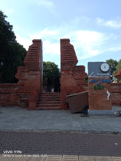 The image shows a brick archway located at the coordinates -6.726, 108.571 in Cirebon, Indonesia. The archway is made of red brick and has a sign above it that reads "masuk gerbang elat" in Indonesian. Two people are walking through the archway. A brown wooden cart is overturned on the ground in front of the archway. The ground is made of gray concrete. The sky is blue with some white clouds.