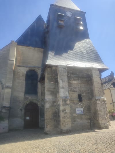 The image shows a close-up, low-angle view of the exterior of the Church of Saint-Rémi in Ressons-sur-Matz, France, on a clear, sunny day. The stone facade of the church, with its weathered texture, is prominent, particularly the large, arched wooden door and a tall, dark, arched window above it. The church features a dark, slate-tiled roof with a steep pitch. A clock tower with a Roman numeral clock face is visible at the upper part of the church. The ground in front of the church is paved with cobblestones. The environment is characterized by a bright blue sky and shadows cast by the sun. No people are visible in the frame.