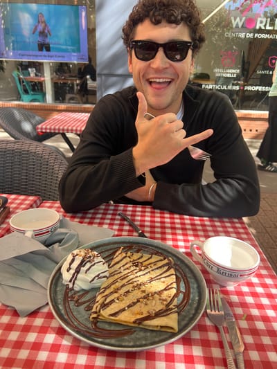 A close-up, eye-level shot captures a male subject seated at an outdoor cafe table, featuring a red and white checkered tablecloth, in Orléans, France. The subject, a man with curly dark hair and wearing black Wayfarer-style sunglasses and a black long-sleeved polo or zippered sweater, grins broadly at the camera with his mouth open and tongue slightly extended. His left hand is raised, making a "shaka" gesture while simultaneously holding a silver fork between his thumb and index finger. A thin gold chain bracelet is visible on his left wrist, and a silver bangle on his right.
In the foreground, on the table, a plate containing a folded crêpe and a dollop of whipped cream is presented. The crêpe is drizzled with dark chocolate sauce and sprinkled with crushed nuts. The whipped cream is similarly adorned with chocolate sauce and nuts. To the left of the plate, a white ceramic bowl with red stripes and "Cidre" printed on it sits empty. To the right, another white ceramic cup with red stripes and "Libre Kerisac" printed on it contains a clear liquid. A fork and knife rest beside this cup.
The background, visible through a glass window, reveals the interior of a business identified as "WORLD CENTRE DE FORMATION," located at 2 Rue Jeanne d'Arc, Orléans, France, based on visible signage. Text on the glass or interior also lists "INTELLIGENCE ARTIFICIELLE," "SANTÉ &," and "PARTICULIERS • PROFESSIONNELS." A large screen inside displays a blonde woman, with text partially visible as "S'ambiancer de Sign Events." Additional outdoor cafe seating with wicker-style chairs and tables covered in red and white checkered tablecloths, along with a brick pavement, are visible in the blurred background. A partially visible figure in dark attire with white striped footwear is on the far right.