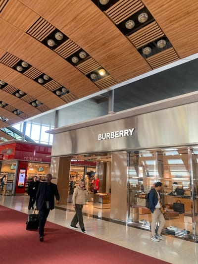 The image captures an indoor commercial concourse, identified as being within Charles de Gaulle Airport in Le Mesnil-Amelot, France. The prominent feature is a Burberry luxury retail store with an illuminated "BURBERRY" sign set against a brushed metallic facade. Its large glass display windows showcase merchandise, including multiple handbags (e.g., a black tote, a brown leather backpack) and various shoes. To the left, a red-fronted store, visible through reflections and direct view, is identified by its interior "RELAY" (reversed) and "Daily" signage, displaying various products and a mannequin in a red jacket.
The ceiling features curved, slatted wooden panels interspersed with numerous circular recessed light fixtures. A bright, light-colored reflective floor extends in front of the stores, met by a wide red carpet pathway that traverses the foreground from left to right. Natural light streams in from large windows positioned in the upper-left background.
Four individuals are present. In the foreground, an adult male of dark complexion, dressed in a dark suit with a gray garment tied at his waist, walks from left to right, pulling a black wheeled suitcase. Behind him, an adult female in a dark blazer is partially visible, also walking. In the mid-ground, an adult female with light-colored hair, wearing a beige top and dark trousers, walks from left to right while speaking on a mobile phone held to her left ear. To the right, an adult male of East Asian descent, wearing a dark blue hoodie, light trousers, and carrying a tan shoulder bag, stands directly in front of the Burberry display window, observing the store's contents. No direct interactions between individuals are discernible.