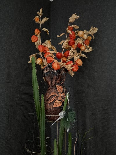 A still life photograph depicts a corner arrangement of decorative elements against a dark, textured wallpaper. At the center, a reddish-brown, ornate ceramic vase, with a floral relief, holds a dried arrangement of Physalis (Chinese lanterns). The lanterns are predominantly orange and faded brown, with dried brown leaves. To the left of the vase, a tall, green cactus with prominent ribs stands in a metal stand. A small, artificial pine sprig with a white ribbon is attached to the metal stand near the vase. The background consists of a dark, patterned wallpaper with a geometric design. The scene appears to be indoors, likely in a domestic setting, in the city of Galinė, Lithuania. No people or animals are visible in the image.