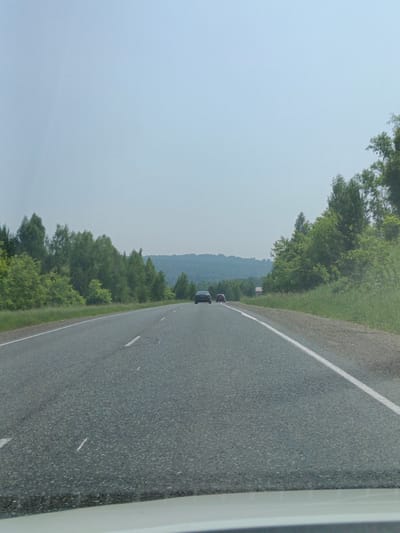The image depicts a view from a vehicle driving on a two-lane asphalt road. The road has white dashed lines indicating lanes. On the left side of the road, there is a dense line of green trees and foliage. On the right side, the vegetation continues, and further in the distance, rolling hills covered in trees are visible. The sky is overcast with a pale blue hue. Two cars are visible in the distance on the road, one dark-colored and one red. The perspective suggests the photo was taken from the driver's or front passenger's seat of a moving vehicle, with the dashboard visible at the bottom of the frame. The overall environment is rural and appears to be a highway or a major road passing through a wooded, hilly landscape.