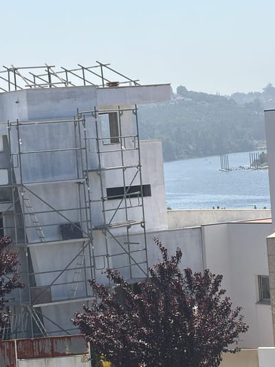 This media file displays a building under construction, likely in Gondomar, Portugal, given the context. The scene is dominated by scaffolding surrounding a partially finished concrete structure. The building appears to be modern, with large, smooth concrete surfaces and rectangular openings for windows. A dark red-leaved tree is visible on the right side, partially obscuring the building. In the background, a body of water, possibly a river or lake, can be seen, bordered by a lush, green hillside with scattered buildings. The sky is clear and pale, suggesting daytime and fair weather. The image is oriented vertically. No people are visible, and the focus is on the construction site and the surrounding landscape. The overall impression is one of ongoing development and a scenic natural setting.