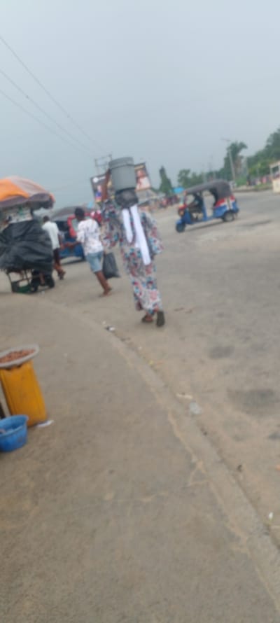 The image depicts a street scene in Yenagoa, Nigeria. In the foreground, a person in a long, patterned dress and white head covering walks away from the camera, carrying a large bucket on their head. They are also holding a black bag in their right hand. To the left, a stall is visible with a yellow container filled with food items, next to a blue bucket. Further in the background, other individuals are present, some near a covered stall with an orange and white umbrella. A blue auto-rickshaw is also seen on the road, with a person driving it. Billboards are visible in the distance, and trees and buildings line the street. The sky is overcast, suggesting a cloudy day. The ground appears to be paved road.