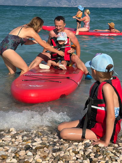 The image depicts a family enjoying a day at the beach in Baška, Croatia. The scene is a waterfront with clear blue water and a pebbly shore. A large, bright red inflatable object, possibly a raft or paddleboard, is central to the composition.
Several people are interacting with the inflatable. A man, appearing to be a father, is holding onto the red object and looking towards a woman. The woman, in a patterned swimsuit, is also engaged with the inflatable. Two children are also present. One child, wearing a life vest and a blue cap, is positioned on the red inflatable. Another child, wearing a yellow and orange top and a blue cap, is next to another woman on a similar red inflatable further in the water. A man in a straw hat is visible on a red inflatable in the background.
The activity appears to be recreational water play, possibly preparing to paddle or simply enjoying the inflatable on the water. The sun is bright, suggesting it is daytime, and the water is calm, indicating pleasant weather. The presence of the inflatable objects and the rocky beach strongly suggests a beach location. The water is shimmering, and small waves are lapping at the shore.
Visible text on the red inflatable reads "MONSTER". The overall emotion conveyed is one of family fun and leisure in a beautiful coastal setting.