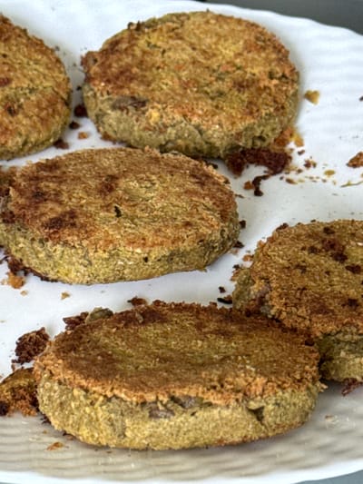 An overhead, close-up shot captures several irregularly shaped, roughly circular to oval food items resting on a white surface, possibly a plate or paper towel. The food items exhibit a greenish-brown interior and are covered with a textured, golden-brown, crispy exterior coating, suggesting they have been fried, baked, or air-fried. Scattered dark brown crumbs are visible on the white surface around and between the pieces. The items vary slightly in size and shape, indicating they may be handmade. They resemble vegetable patties, falafel, or fried cutlets. No activity is explicitly shown, but the food appears to be prepared and ready to be eaten. There is no visible text, and the time of day, weather, and specific location cues within the image are not discernible.