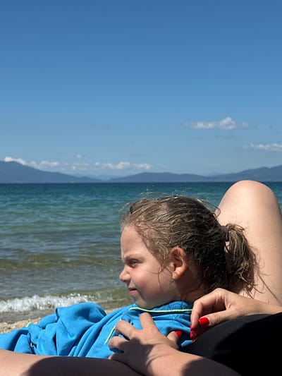 A close-up shot shows a young girl lying on a beach, with her head resting on an adult's lap. The girl is wearing a blue hooded jacket, and her hair is wet and pulled back into a ponytail. Her eyes are closed, and she appears to be relaxed. The adult's hands are visible, with bright red nail polish. In the background, a calm turquoise sea meets a distant mountainous horizon under a clear blue sky. The image is taken from a low angle, with the focus on the girl and the adult's legs. The scene suggests a peaceful day at the beach, possibly in the city of Ofrynio Beach, Greece.