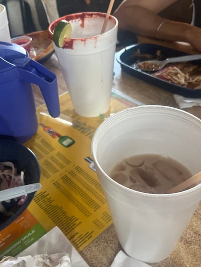 A cluttered table presents a casual dining scene. Styrofoam cups hold beverages; one, a brown liquid with ice, the other rimmed with red and garnished with lime. A blue water dispenser sits nearby. A dark dish filled with shredded food rests close to a menu card with densely printed text. Another dark dish contains what appears to be a salad of shredded cabbage and onion. A person's arm is visible in the upper left corner, and crumpled napkins complete the scene.