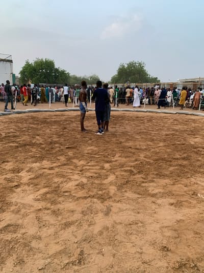 The image shows a dirt arena with a group of people standing around it. There are three men in the center of the arena, and a larger crowd of men are standing around the edge of the arena. There is also a white building in the background and a few trees. The location is Sokoto, Nigeria, at the coordinates 13.049 latitude and 5.238 longitude.