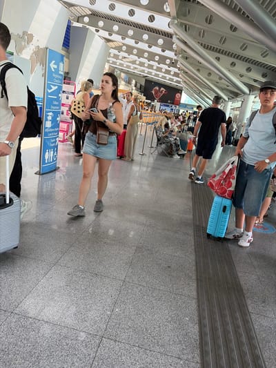 The image depicts an indoor scene, likely an airport or transportation hub, with a modern architectural design featuring a high ceiling with exposed metal beams and circular lights. The floor is a polished gray speckled tile. Several individuals are present, some walking and others seated. In the foreground, a woman in a denim skirt and brown top is holding a smartphone and a water bottle. To her left, a person is walking with a silver suitcase. To the right, a person wearing a light blue t-shirt and denim shorts is holding a red and white floral bag on top of a blue suitcase. A directional signpost in the background indicates "Registration Check-in", "Odlasci Departures", and "Izgubljeno Nađeno Lost & Found". The overall impression is one of transit and travel within a public facility. The location is identified as Golubovci, Montenegro.