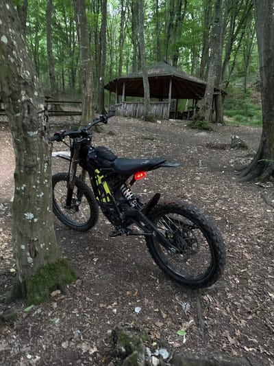 The image depicts an outdoor scene in a wooded area, with a dirt path and a small gazebo in the background. A black electric dirt bike is parked on the path, leaning against a tree. The bike has yellow accents and a red taillight. The environment appears to be a forest with a thick canopy of trees and leaf-covered ground. There is no indication of Kichevo, Bulgaria, or any people present in the image.