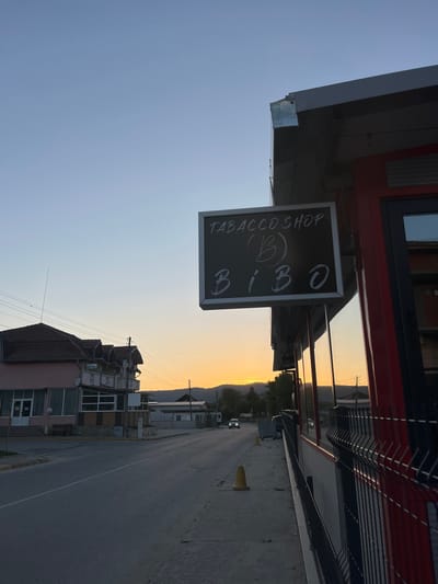 A street scene in Satovcha, Bulgaria, shows a "Tabacco Shop (B) Bibo" sign hanging under the awning of a red-trimmed building. The view looks down a long, straight road with a low mountain range visible in the distance under a pale yellow sky. Buildings line the left side of the street, including a building labeled "Hotel." A single yellow traffic cone sits on the road's edge. A black metal fence runs along the right side of the street.