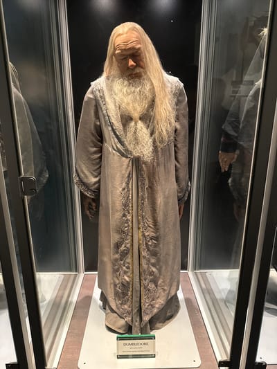 This media file shows a display case containing a wax figure of Albus Dumbledore from Harry Potter. The figure is dressed in a long, grey robe with intricate gold embroidery along the hem and sleeves. Dumbledore has long, flowing blonde hair and a beard, and his face is rendered with aged, wrinkled skin. His right hand is visible, extended slightly from his robe.
The wax figure is positioned in a glass display case, with reflections of other parts of the exhibition visible on the glass. A small plaque is attached to the display, with the visible text reading: "DUMBLEDORE 1:3 scale model Harry Potter and the Half-Blood Prince."
The lighting in the display case appears to be artificial, creating a theatrical effect. The background behind the figure is dark, further highlighting the subject. The setting suggests a museum or exhibition dedicated to the Harry Potter franchise, possibly located in Three Rivers, United Kingdom. No specific time of day or weather conditions are discernible from the image. The figure itself is presented in a static, dignified pose, consistent with a museum exhibit.