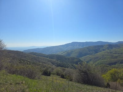 The image depicts a mountainous landscape in the region of Yavrovo, Bulgaria. A clear, blue sky dominates the upper portion of the frame, with a bright sun visible near the top. The landscape consists of layers of hills and mountains covered in green vegetation, indicating a forested area. In the foreground, there's a grassy slope with shrubs and small trees. A winding road is visible in the valley below. In the distance, additional mountain ranges are discernible, fading into a bluish haze towards the horizon.